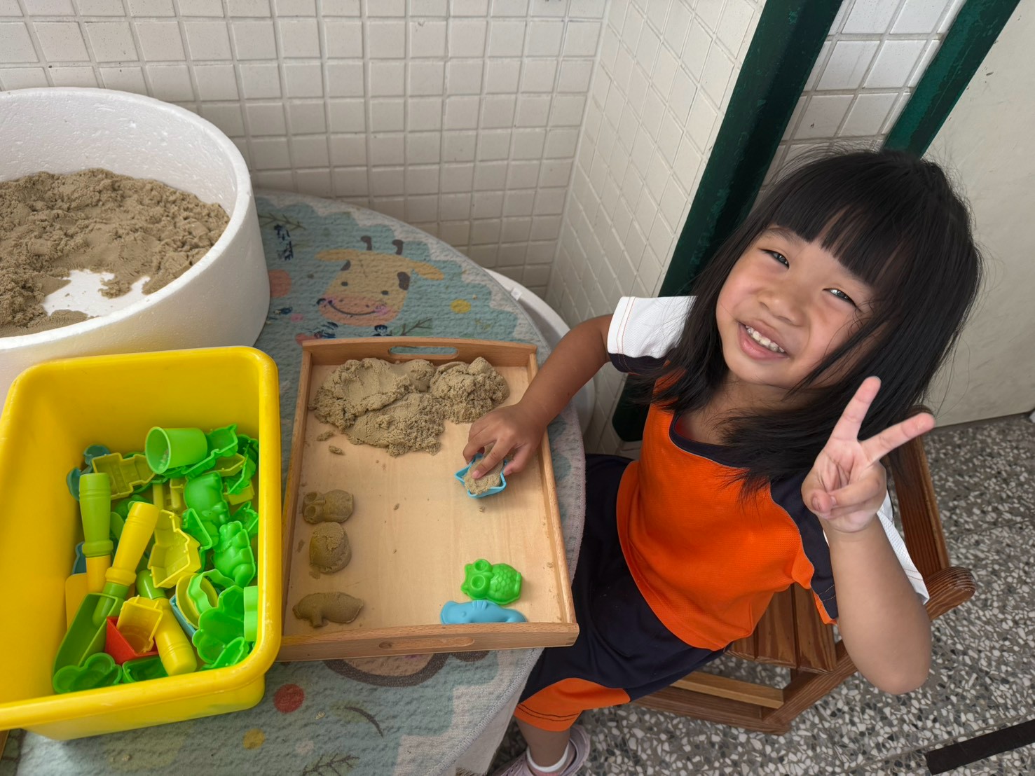 女孩在玩裝在白色大盆與木托盤裡的動力沙/沙土與塑膠模具，這常被規劃在戶外或半戶外的感官探究區（沙水區）。