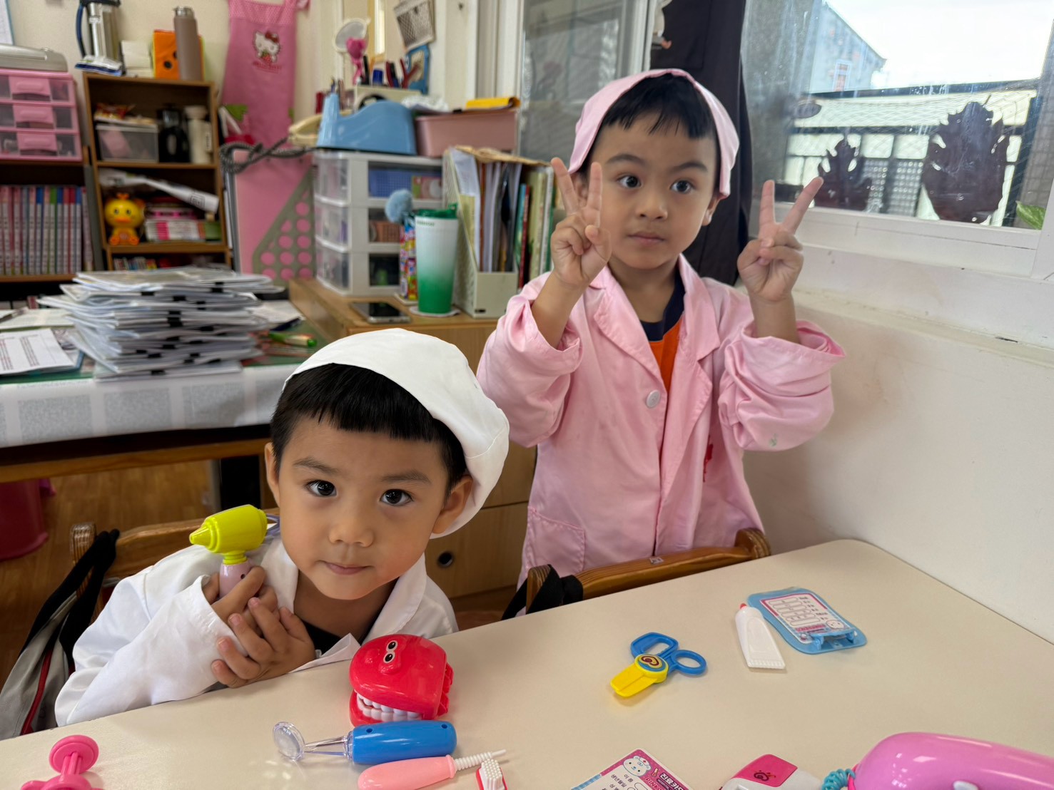 孩子們穿著粉紅色與白色的醫生、護士袍，桌上有牙齒模型、聽診器等工具，正在進行醫院主題的角色扮演。